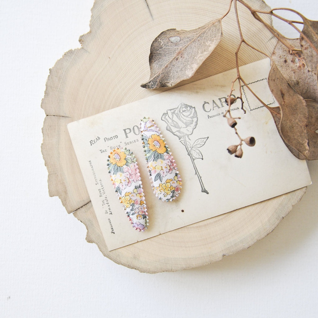 US stockist of Josie Joan's Audrey fabric hair clips.  Set of two fabric hair clips made from pale coloured fabric with bright orange, purple, red, yellow or pink flowers and  contrasting green leaves.  Has scalloped edging.