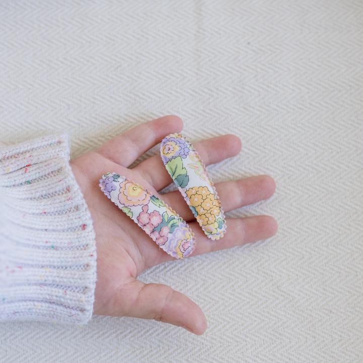 US stockist of Josie Joan's Little Audrey fabric baby hair clips.  Set of two fabric hair clips made from pale coloured fabric with bright orange, purple, red, yellow or pink flowers and  contrasting green leaves.  Has scalloped edging.