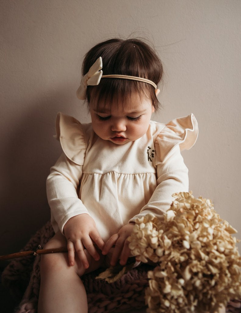 US stockist of Karibou Kid's Milana Winged Playsuit in almond cream.  Made from soft cotton, featuring long sleeves and ruffled wings at shoulders.